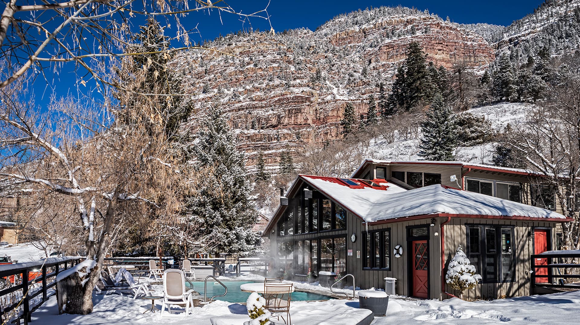 A cozy cabin surrounded by snow-covered mountains, with a steaming hot tub and lounge chairs in the foreground.