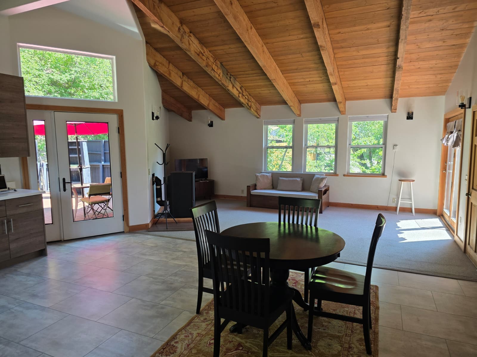 A modern living space with high wooden beams, a dining table, and large windows showcasing greenery outside.