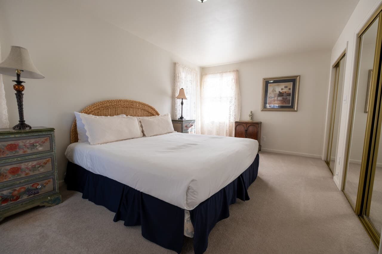 A bright bedroom featuring a large bed with white linens, a wicker headboard, and decor including a lamp and framed artwork.