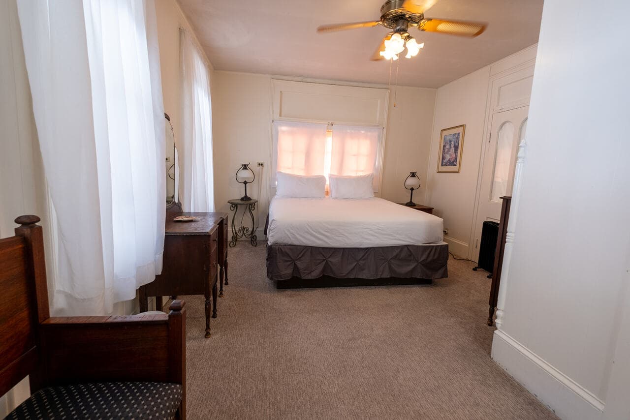A cozy bedroom featuring a double bed, wooden furniture, and large windows with sheer curtains.