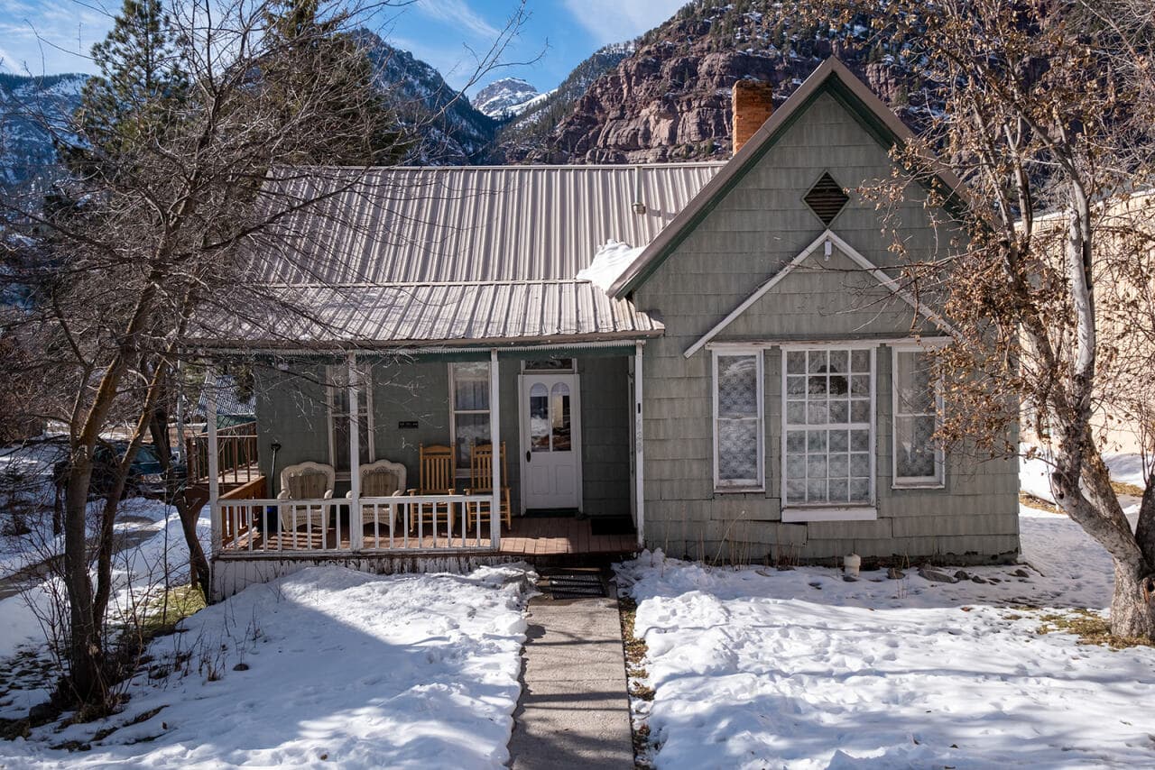 A quaint, snow-covered house with a front porch and surrounded by trees, set against a mountainous backdrop.
