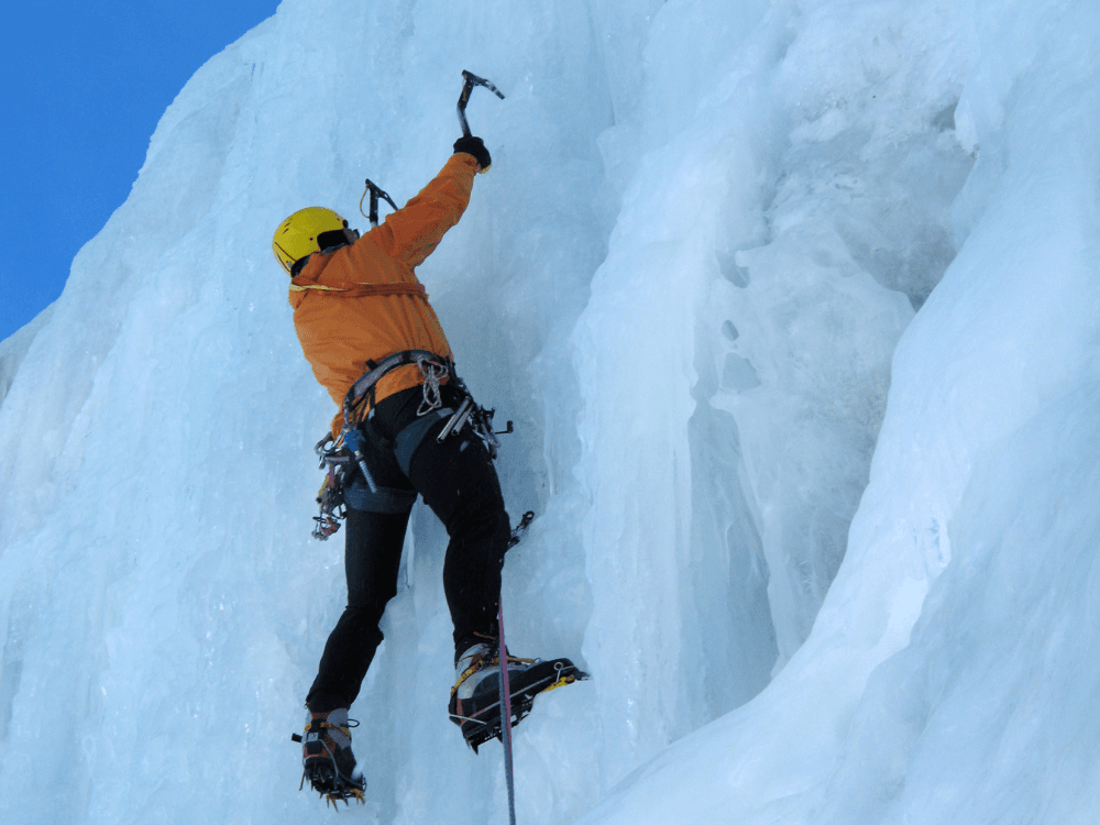 A climber ascends a steep, icy surface using ice axes and crampons.