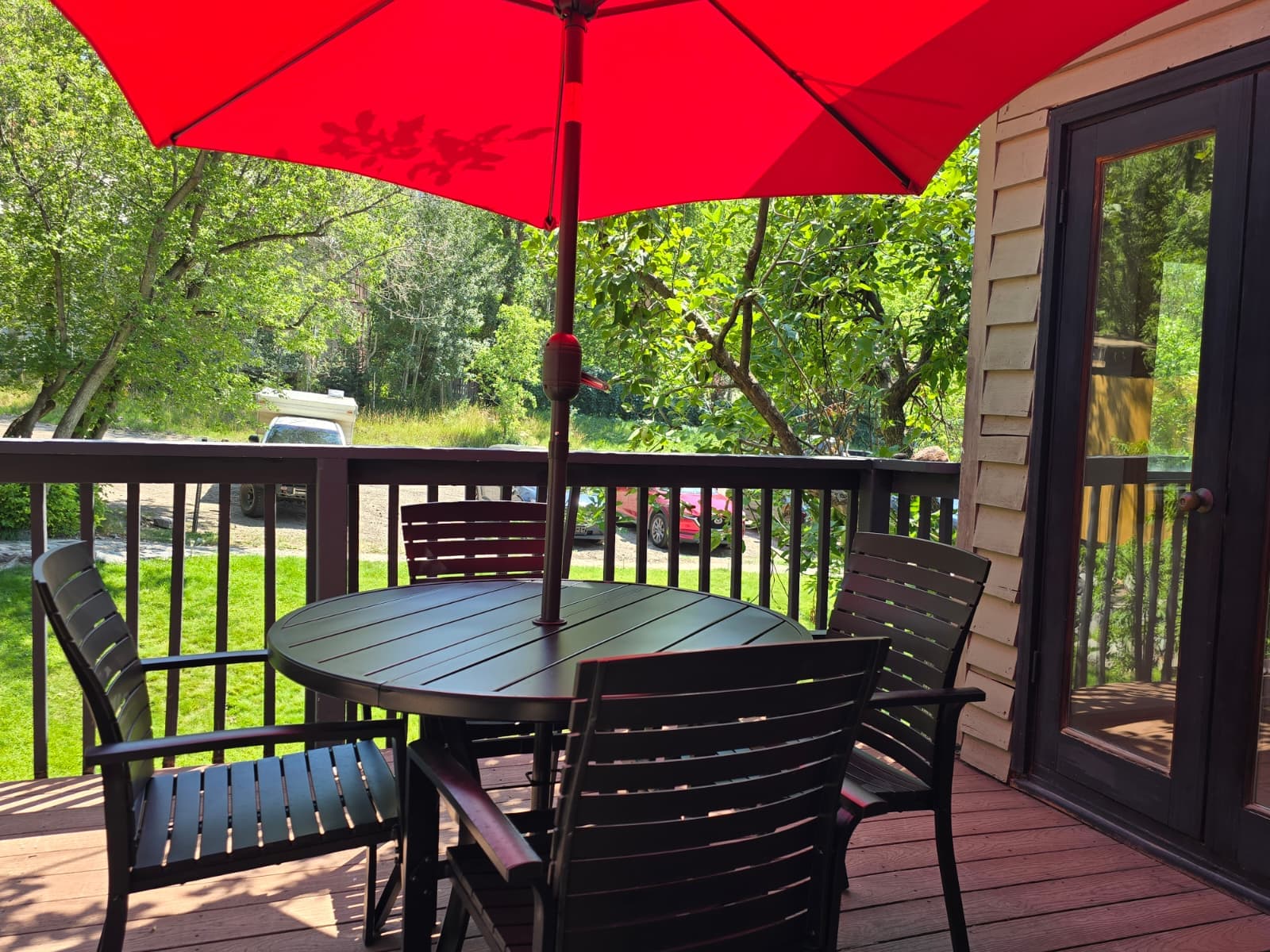 A patio with a round table and black chairs, shaded by a vibrant red umbrella, overlooking a green landscape.