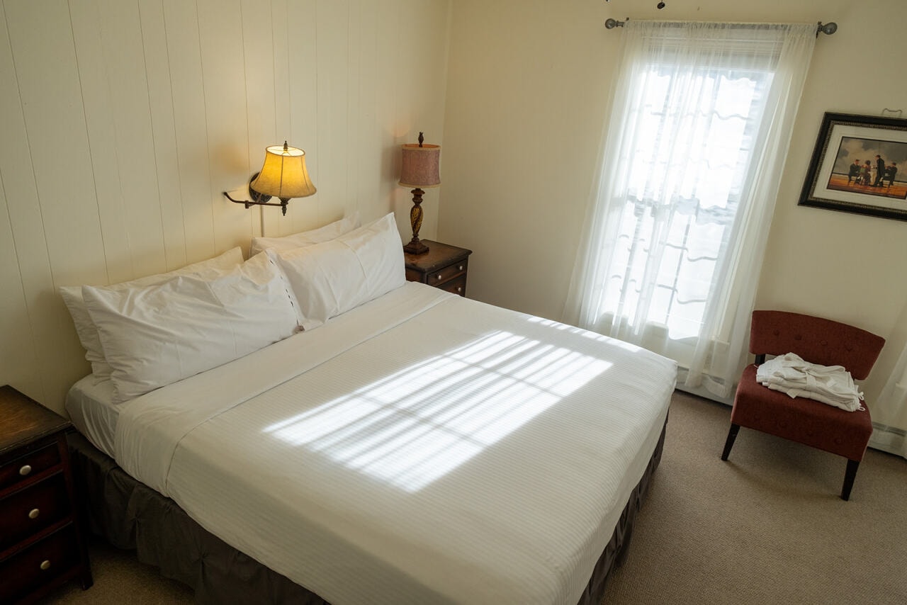 A cozy bedroom featuring a queen-sized bed, a bedside lamp, and a window with soft curtains.