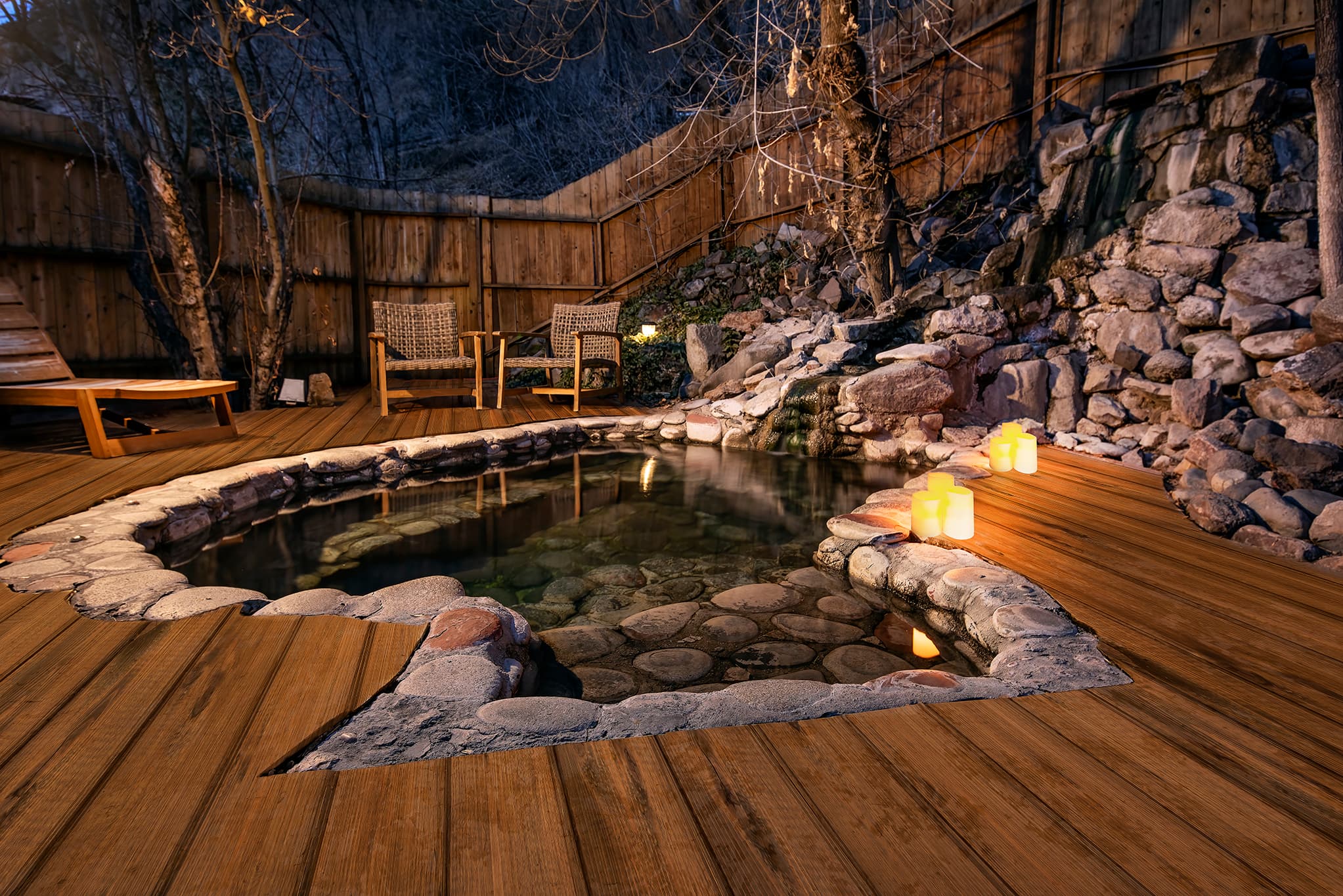 A heart-shaped hot tub surrounded by wooden decking, chairs, and candles in a serene outdoor setting at dusk.