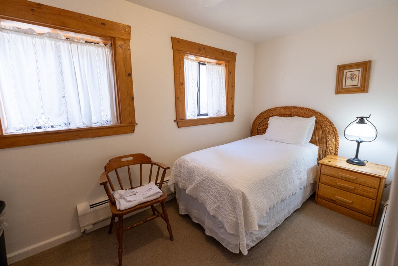A small, cozy bedroom featuring a single bed, wooden furniture, and bright natural light from two windows.