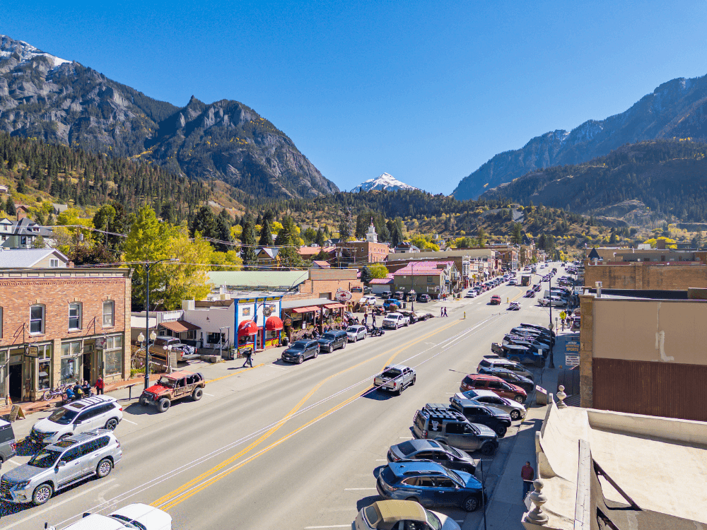 A vibrant town street lined with shops and parked cars, set against a backdrop of mountains under a clear blue sky.