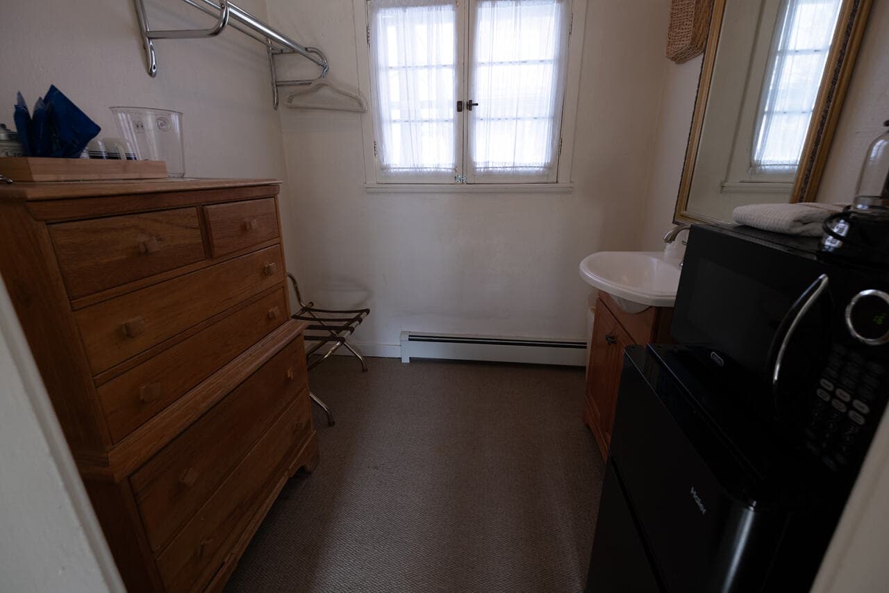 A small, simple room featuring a dresser, mirror, sink, mini-fridge, and a window with sheer curtains.