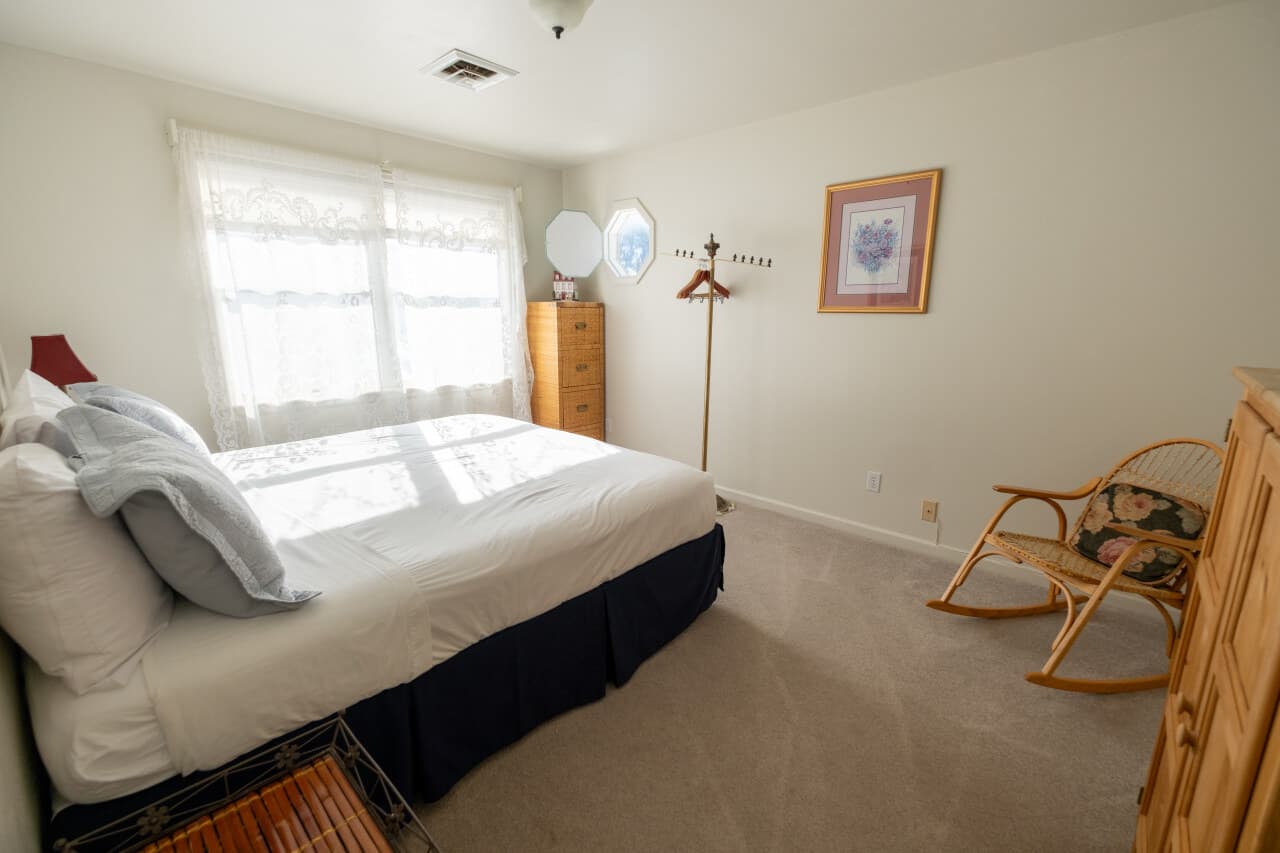 A bright, tidy bedroom featuring a bed, dresser, rocking chair, and a hanger stand by the window.