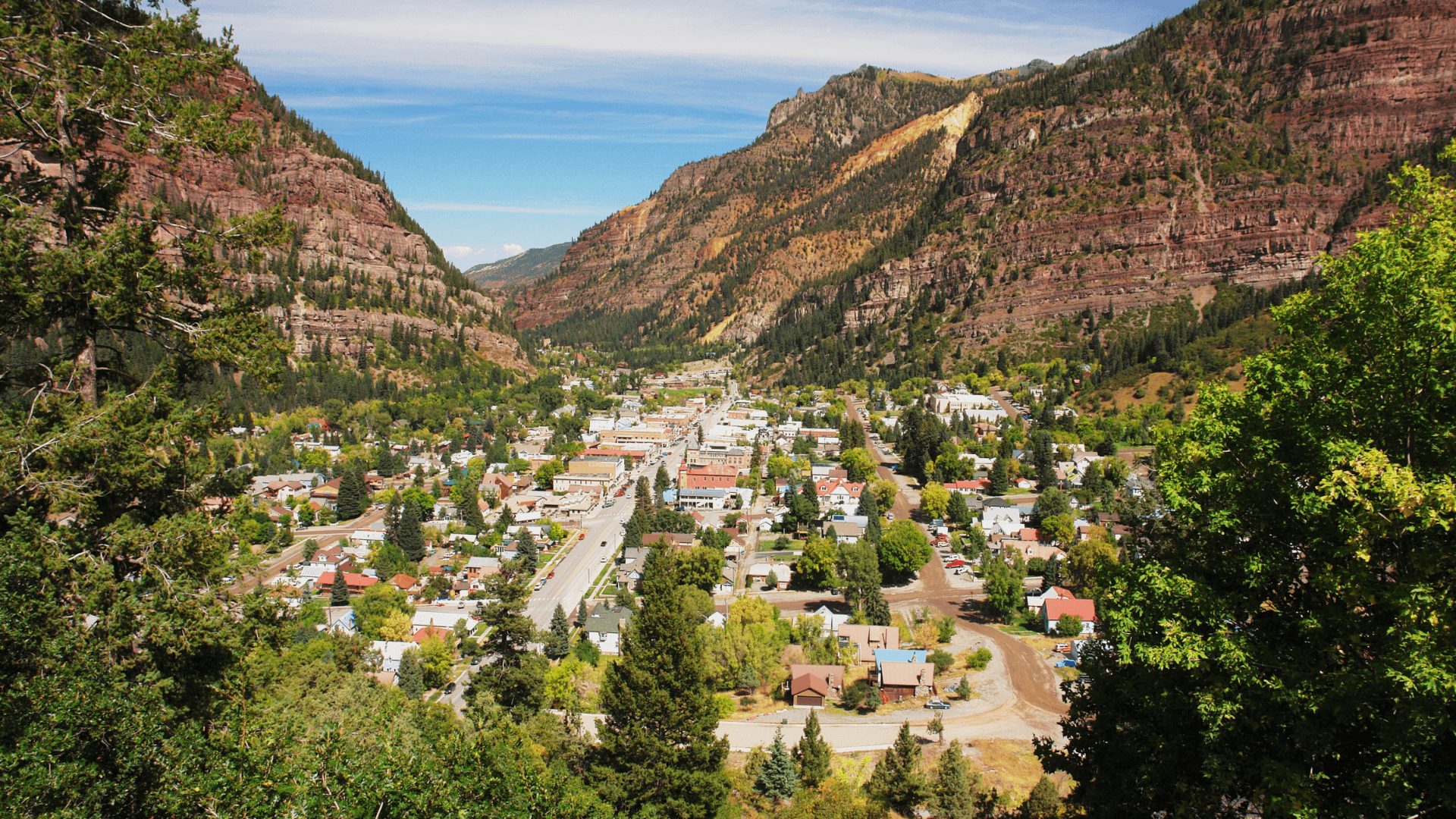 A picturesque valley town surrounded by steep, colorful mountain cliffs under a clear blue sky.