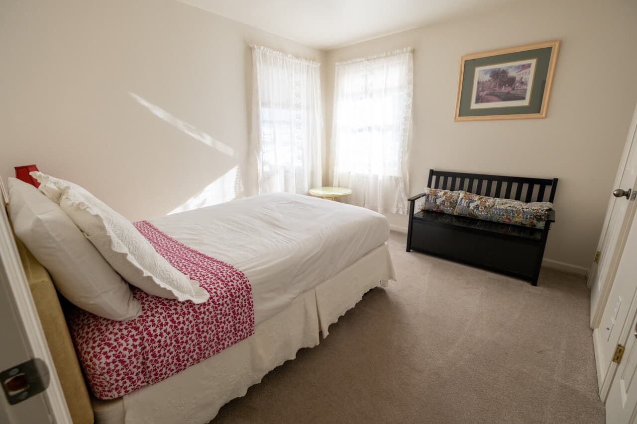 A bright, minimalist bedroom with a twin bed, light curtains, and a small bench against the wall.