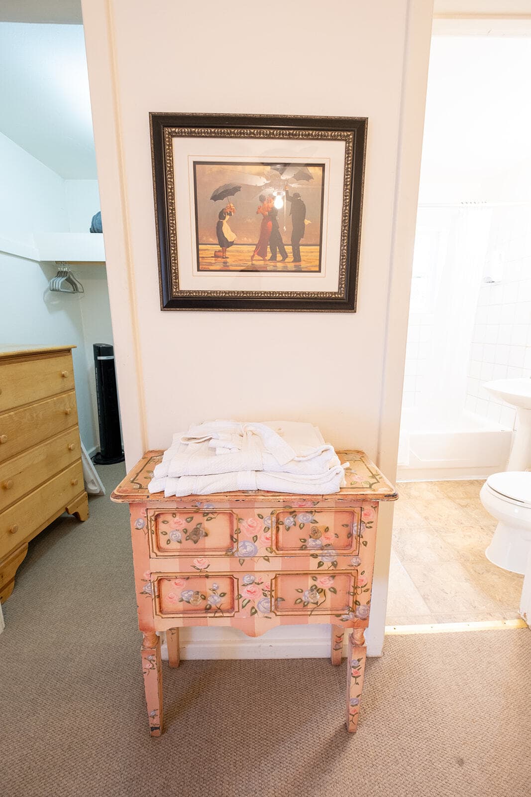 A small, floral-painted chest of drawers topped with folded white towels, beside a framed artwork and a bathroom doorway.