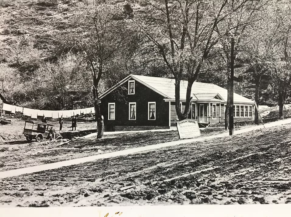 Black and white photograph of a house with a sign, surrounded by trees and laundry hanging outside.