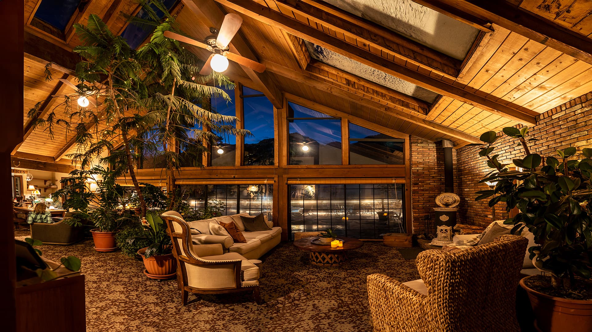 Cozy indoor lounge with wooden beams, large windows, and lush plants, illuminated by soft lighting at dusk.