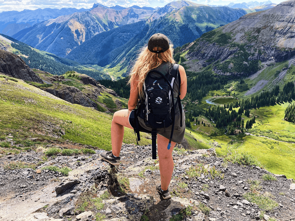 A person with a backpack stands on a rocky outcrop overlooking a lush green valley and mountains.