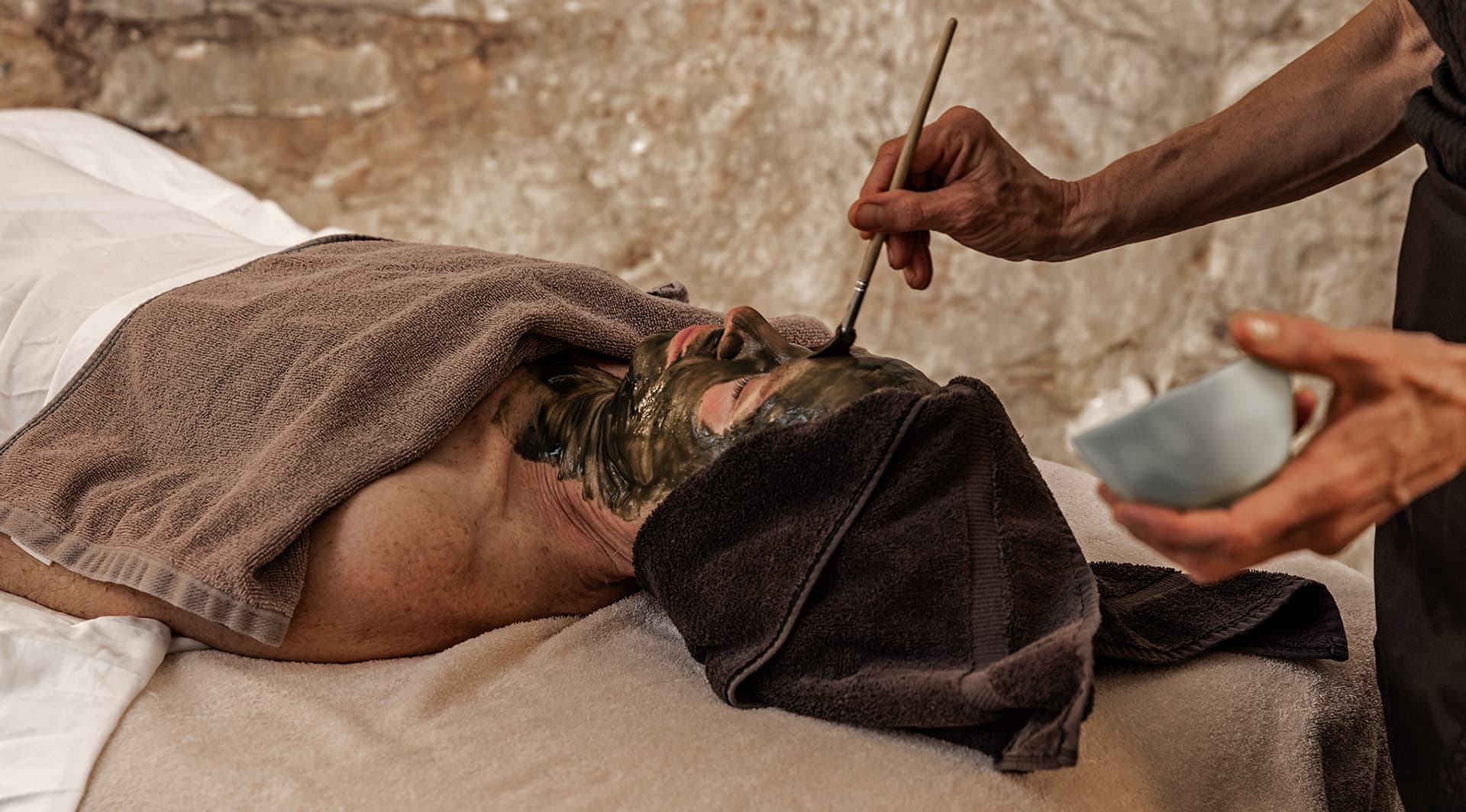 A person receives a facial treatment with a green mask applied while lying on a treatment table.