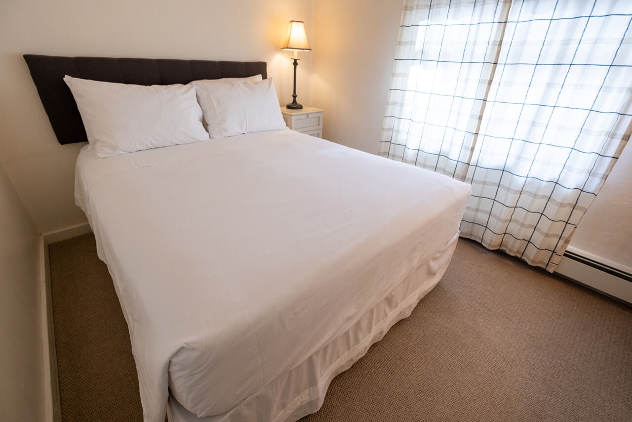 Cozy bedroom featuring a neatly made bed, a bedside lamp, and a window with light curtains.