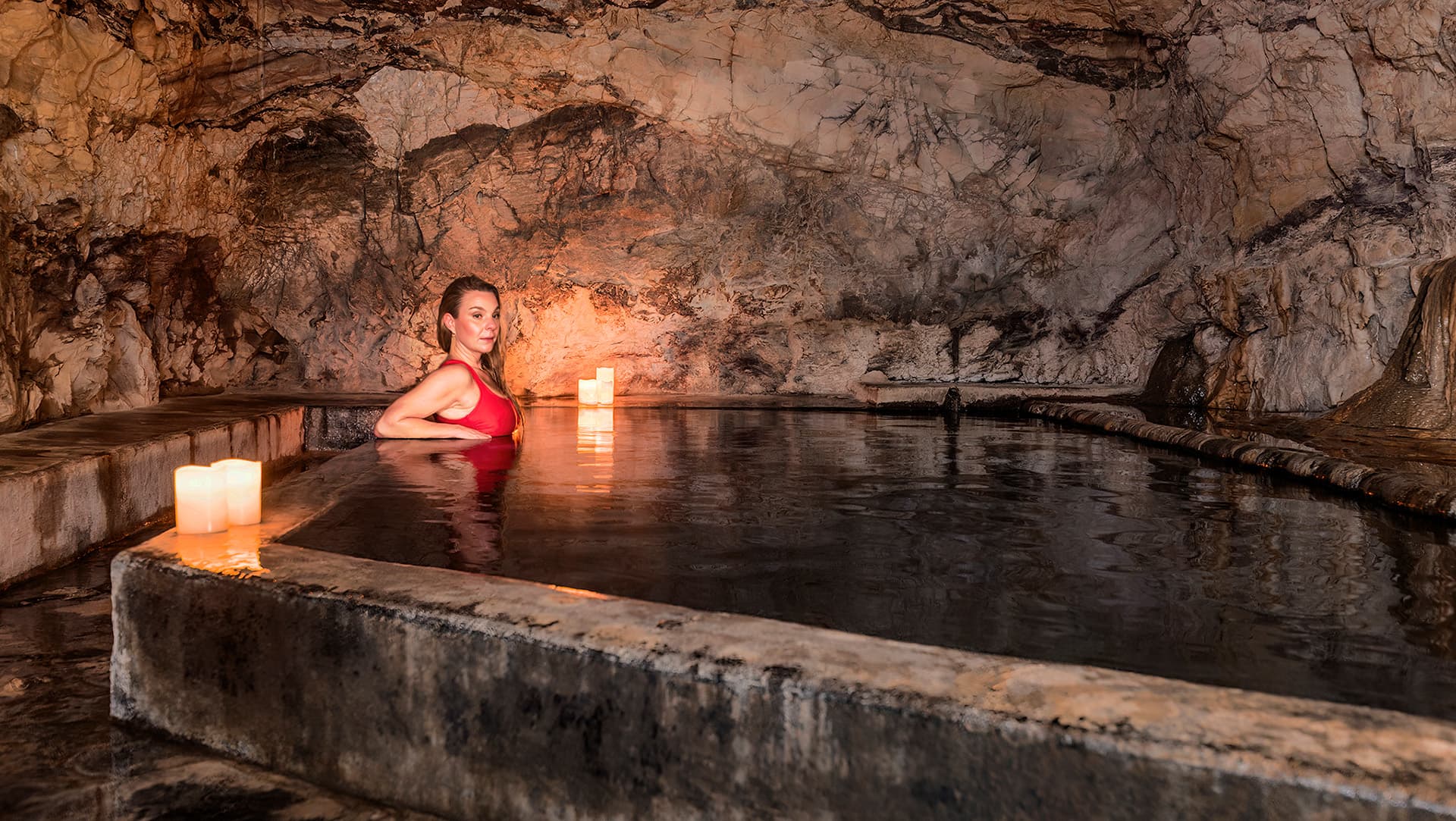 A woman in a red swimsuit relaxes in a dimly lit cave pool surrounded by candles.
