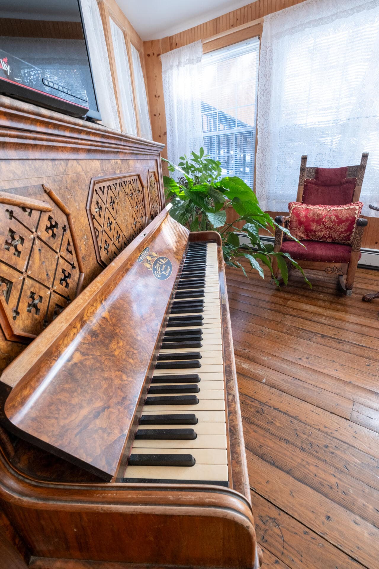 A wooden piano with a decorative design sits beside a lush plant and a cozy armchair in a well-lit room.