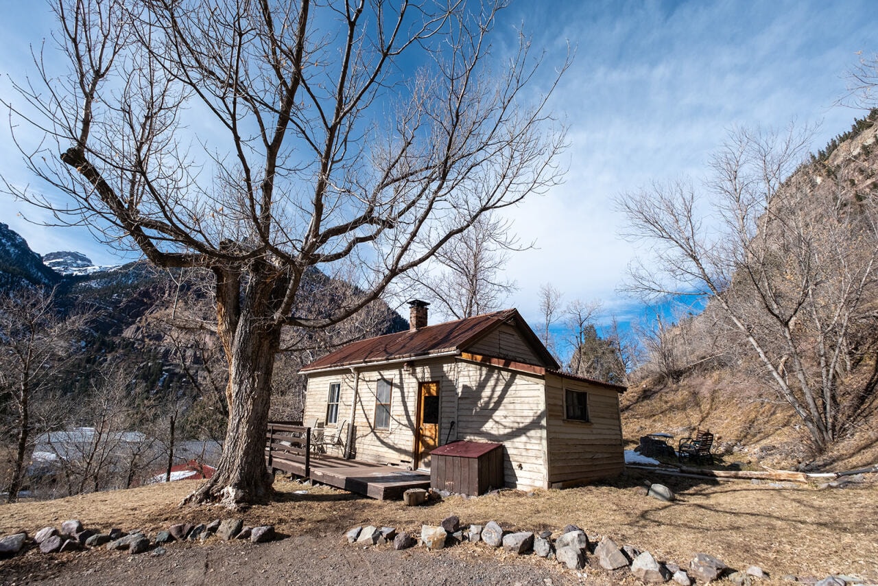 A rustic wooden house sits near a barren tree against a backdrop of mountains and blue sky.