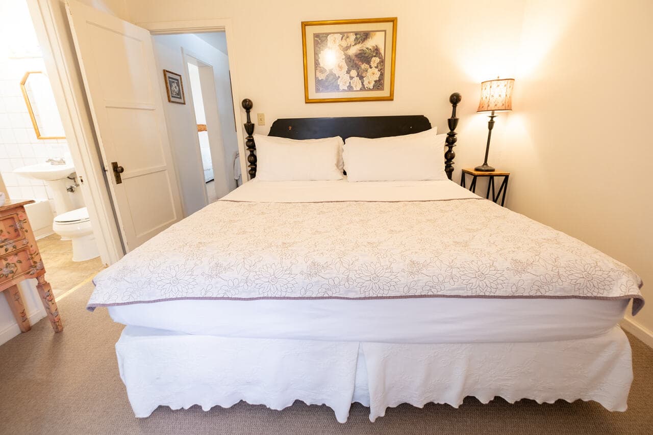 A cozy bedroom featuring a double bed with white linens, a bedside lamp, and a glimpse of an adjacent bathroom.