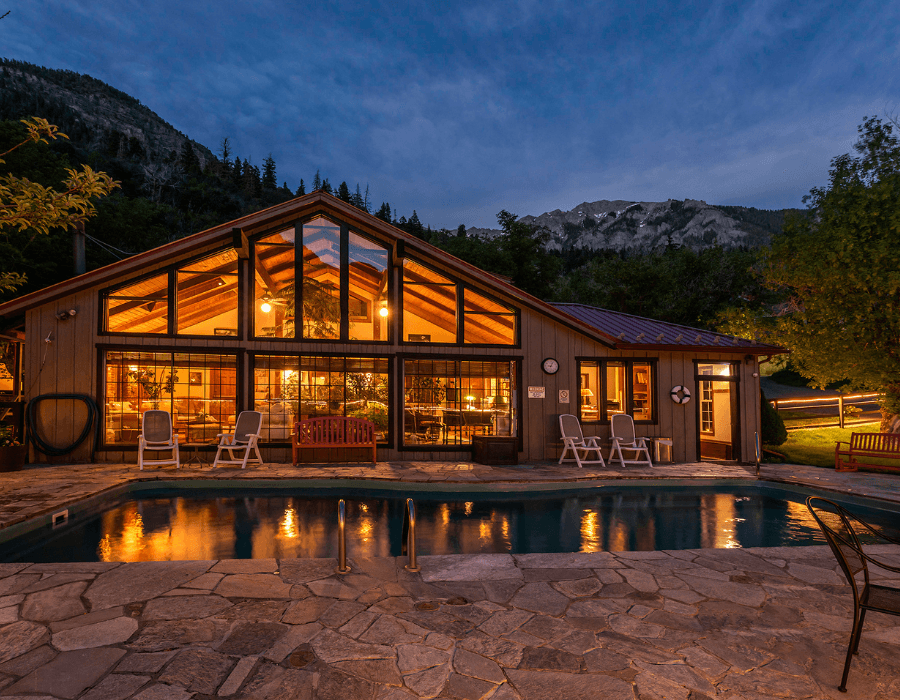 A modern lodge with large windows overlooks a tranquil pool and mountain backdrop at dusk.