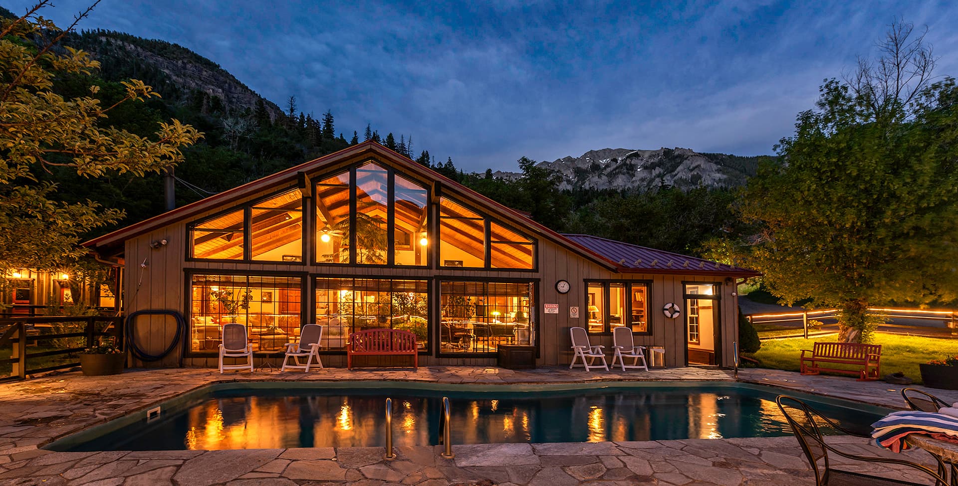 A cozy lodge with large windows and a swimming pool, set against a mountain backdrop at dusk.