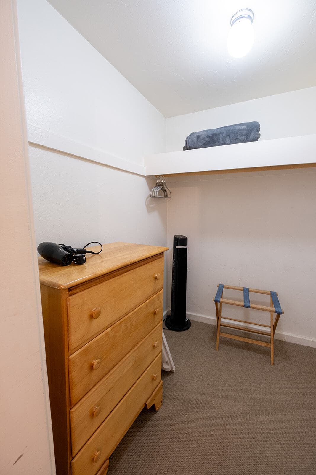 A small, minimalist room featuring a wooden dresser, a folding stool, and a lamp.