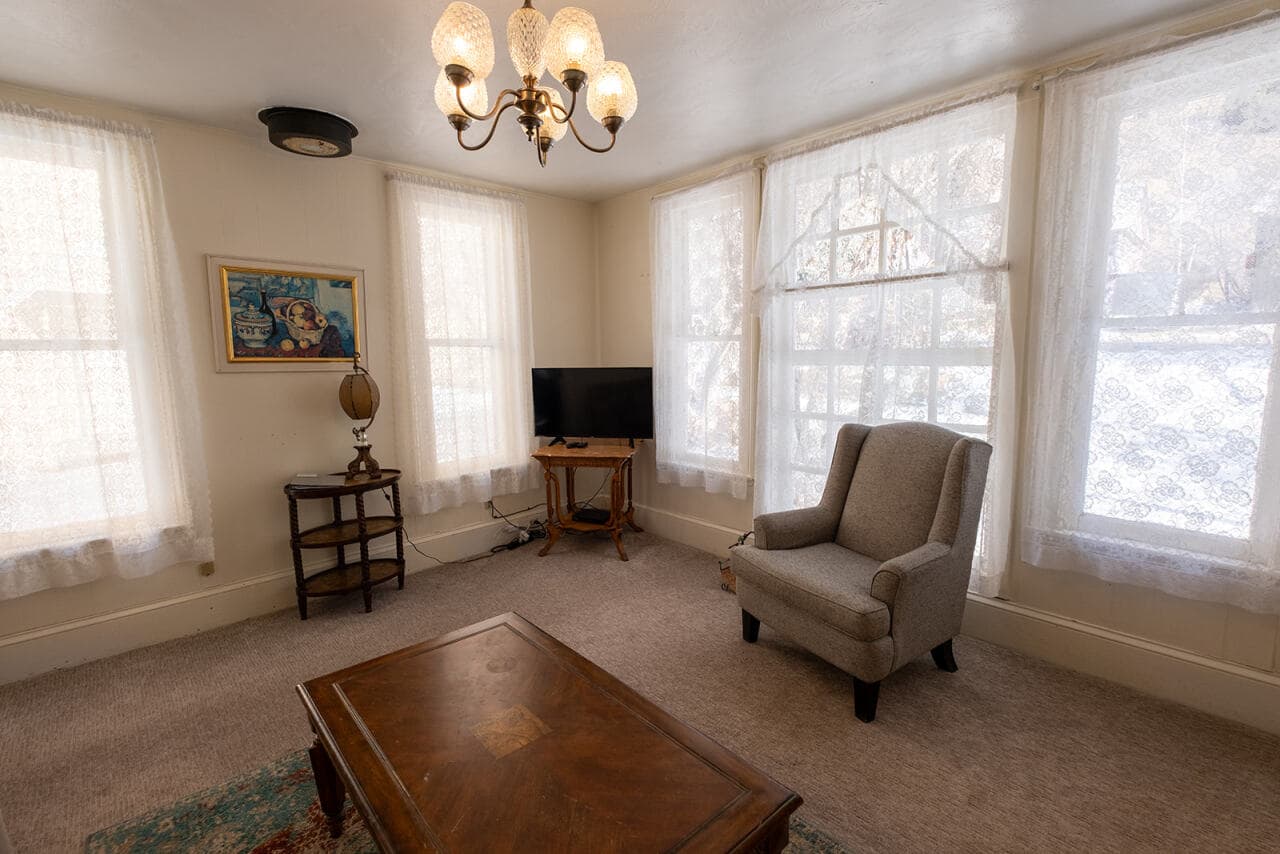 Cozy living room with a gray armchair, a wooden table, a TV, and bright windows.