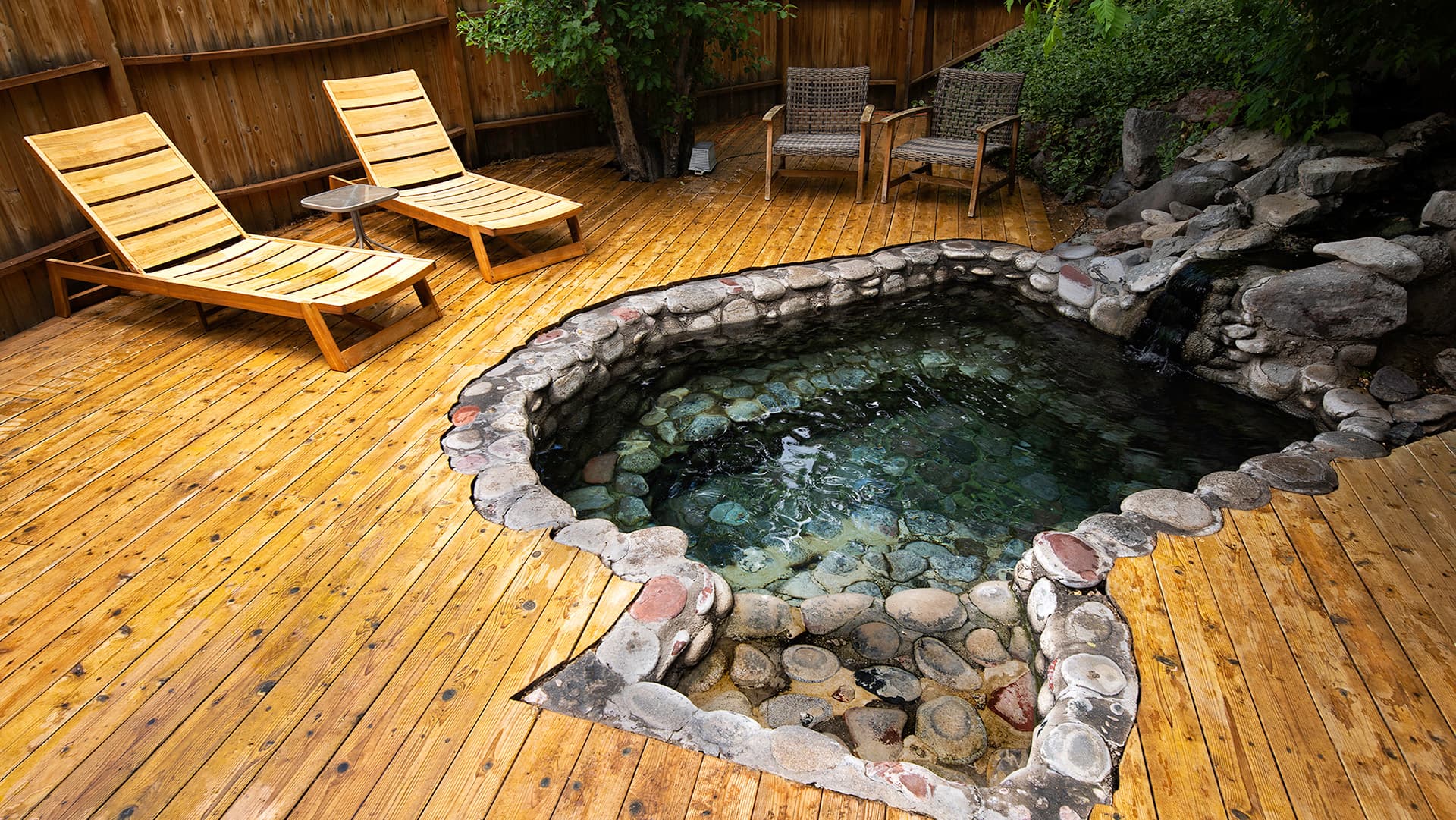A wooden deck features two lounge chairs beside a stone-lined, clear water hot tub.