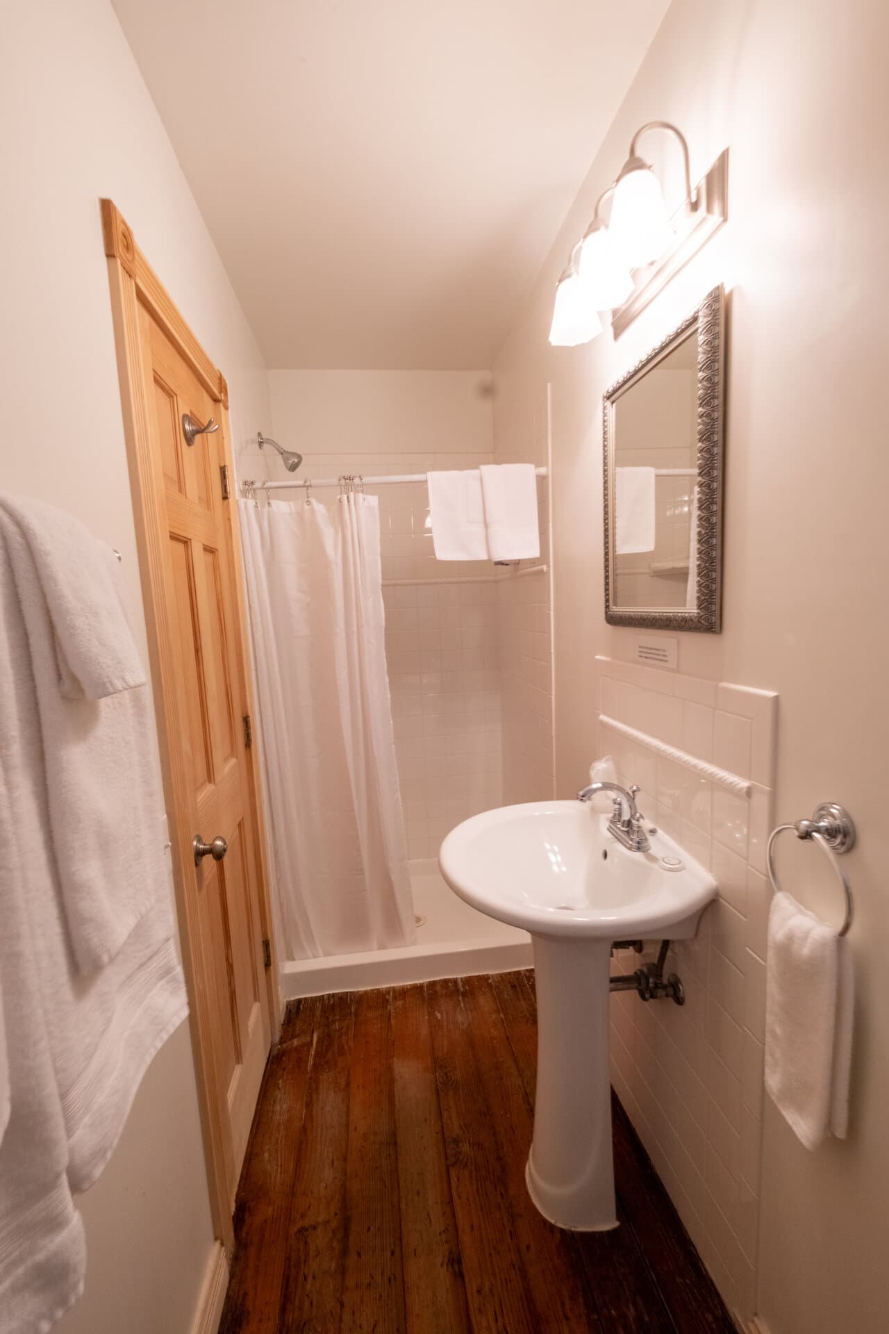 A small bathroom featuring a shower, pedestal sink, and wooden flooring.