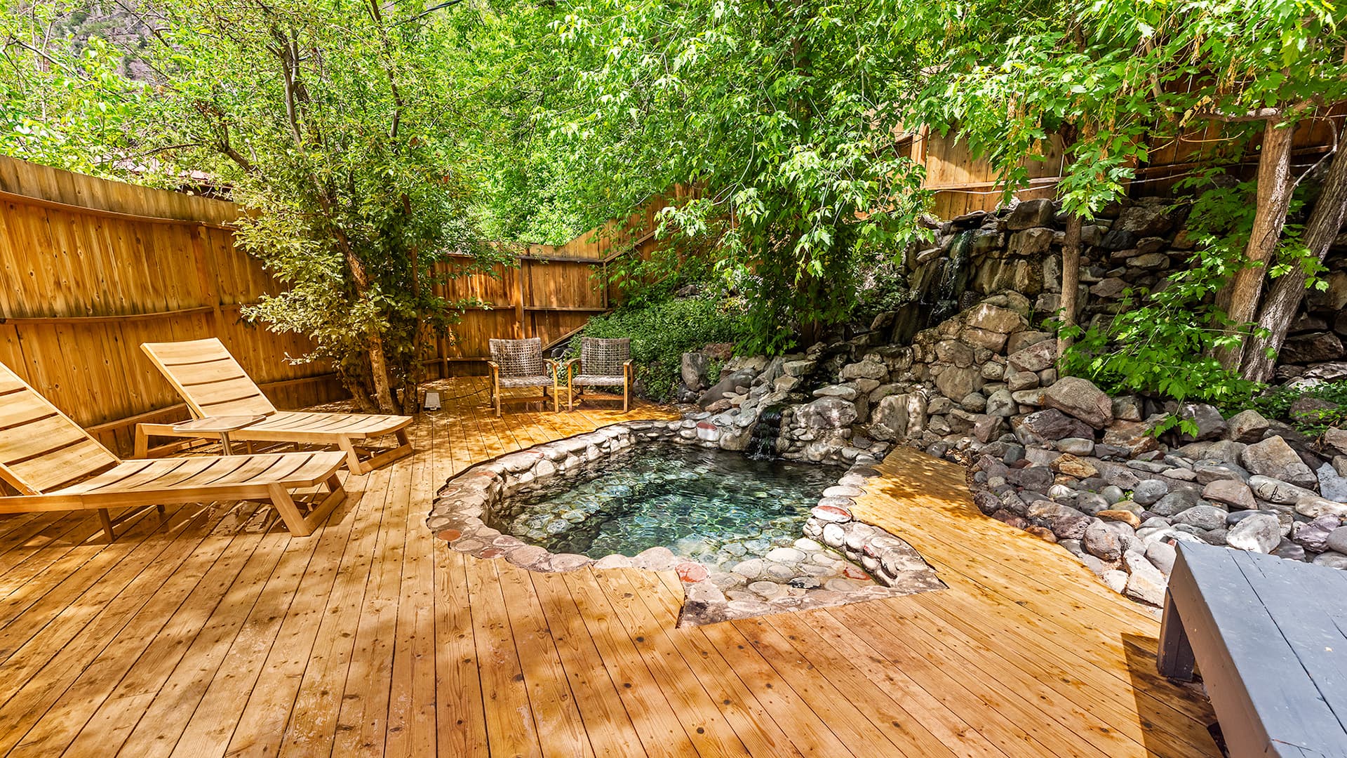 A wooden deck features two lounge chairs beside a natural stone hot tub surrounded by greenery.