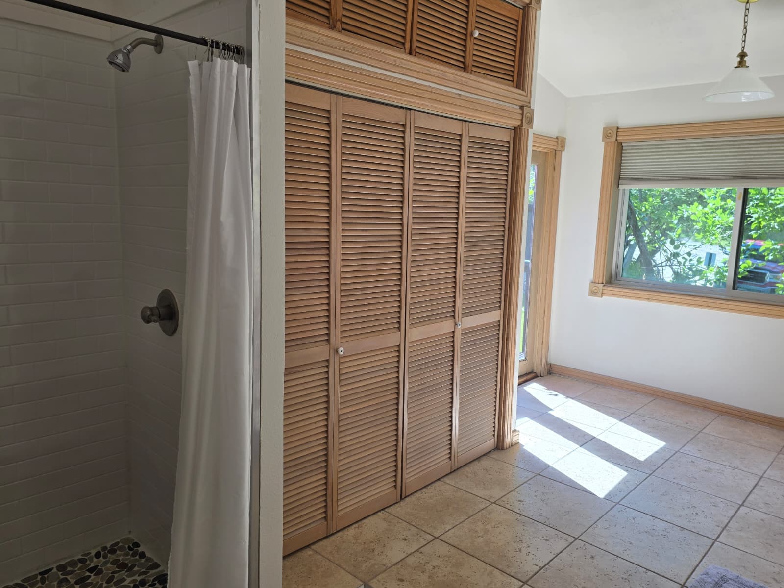 A bright, tiled room featuring a shower area on the left and a wooden closet door on the right, with sunlight streaming through a window.