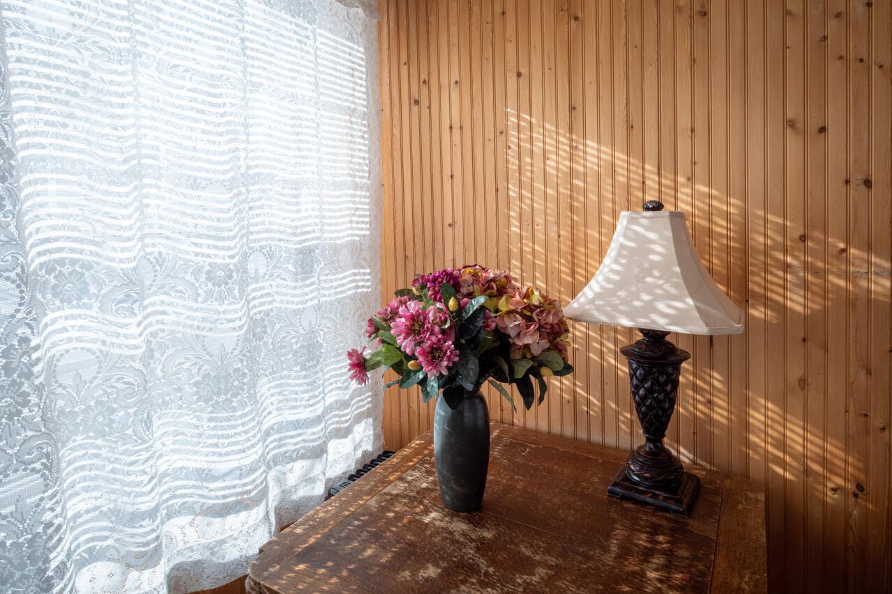 A vase of flowers and a lamp on a wooden table beside a lace curtain.
