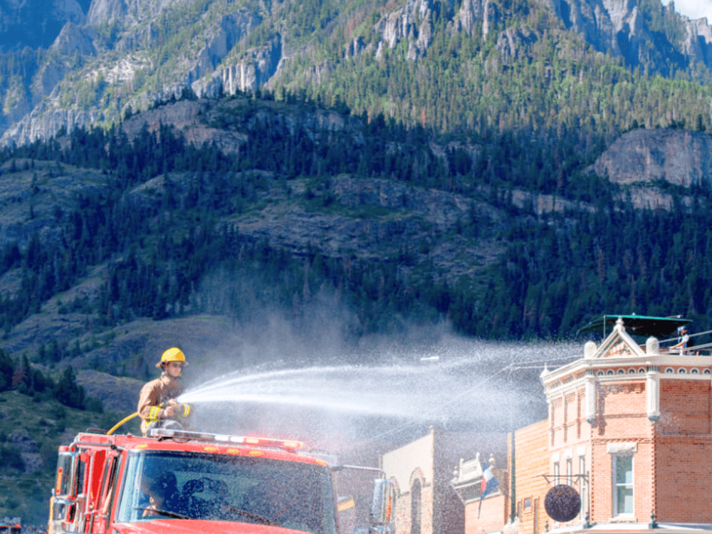 A firefighter sprays water from a fire truck in front of a mountain backdrop.