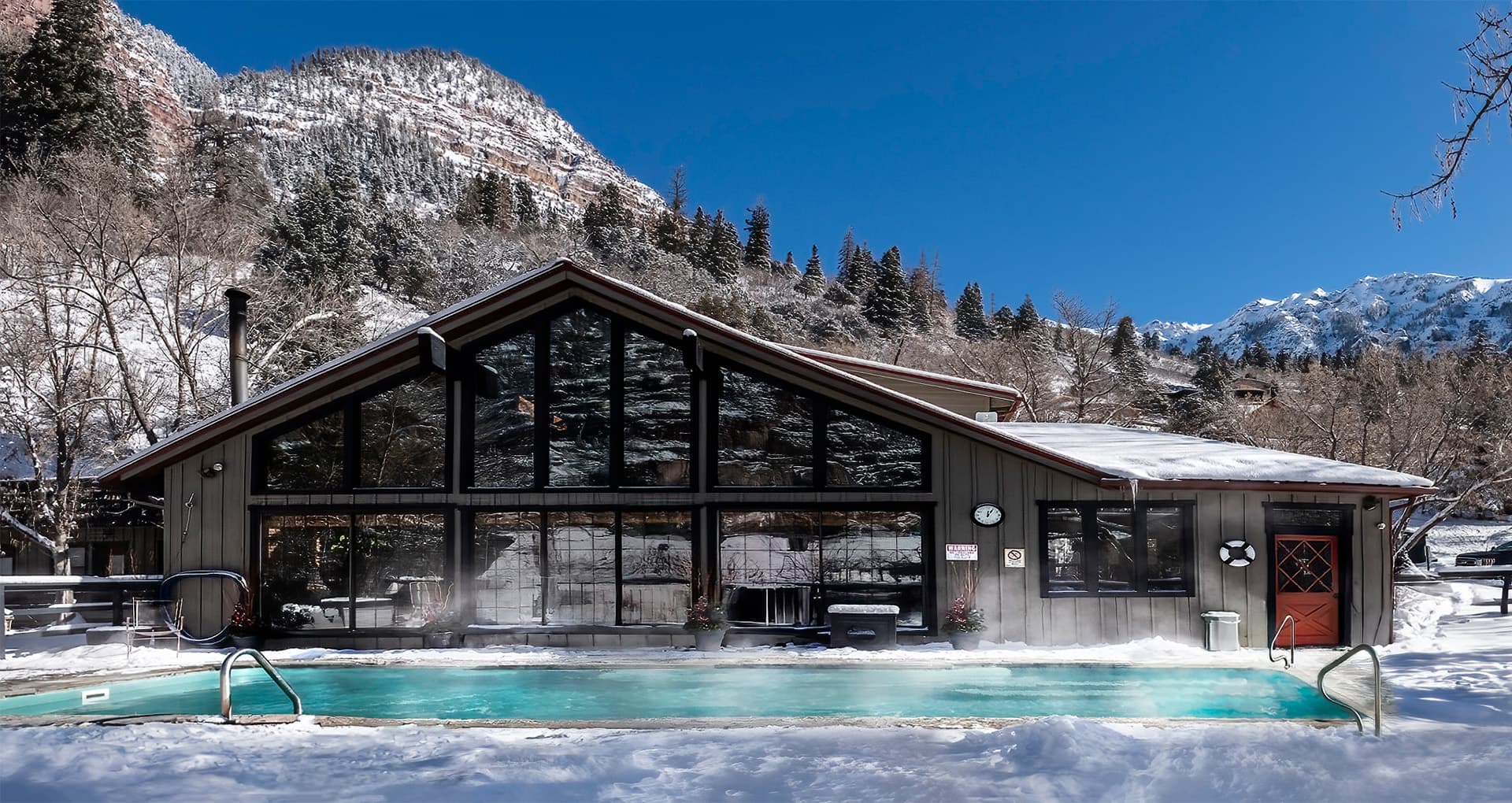A modern building with large windows overlooks a steaming outdoor pool surrounded by snow-covered mountains.