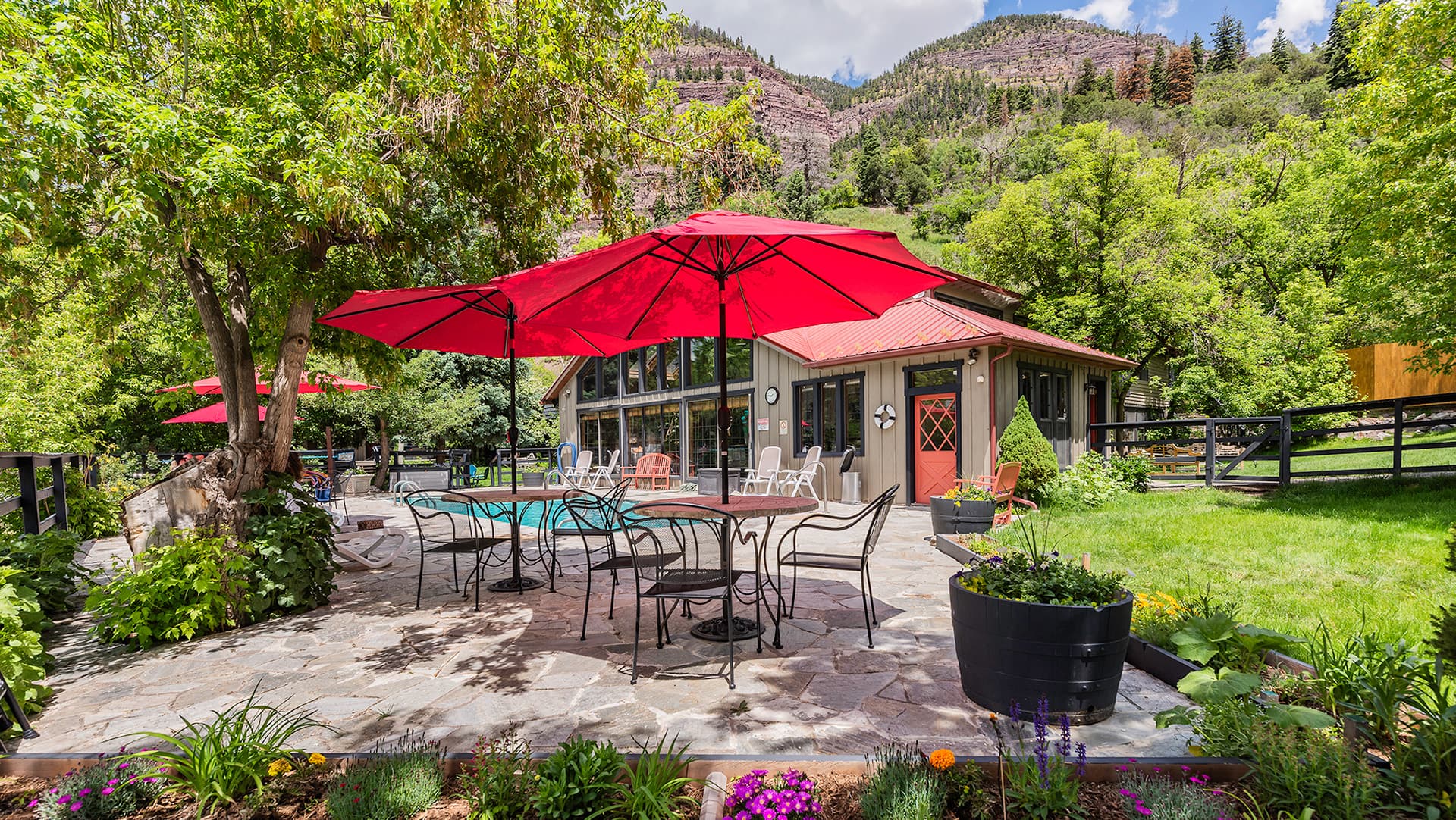 A landscaped patio area with red umbrellas and a rustic building surrounded by trees and mountains.