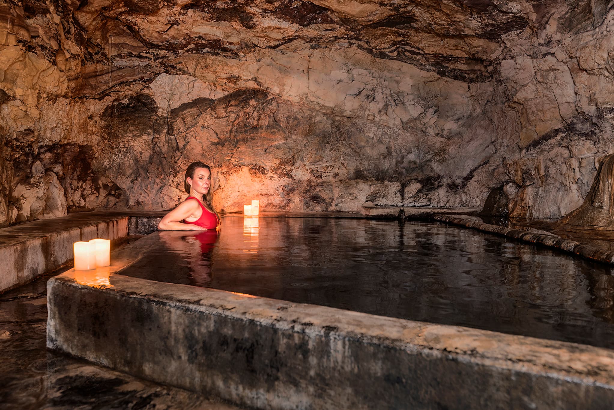 A woman in a red swimsuit relaxes in a dimly lit stone cave with a mineral pool and candles.