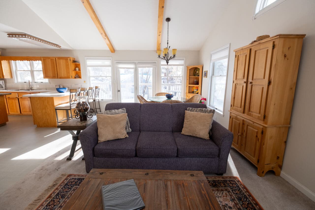 Cozy living room with a purple sofa, wooden furniture, and an open kitchen area bathed in natural light.
